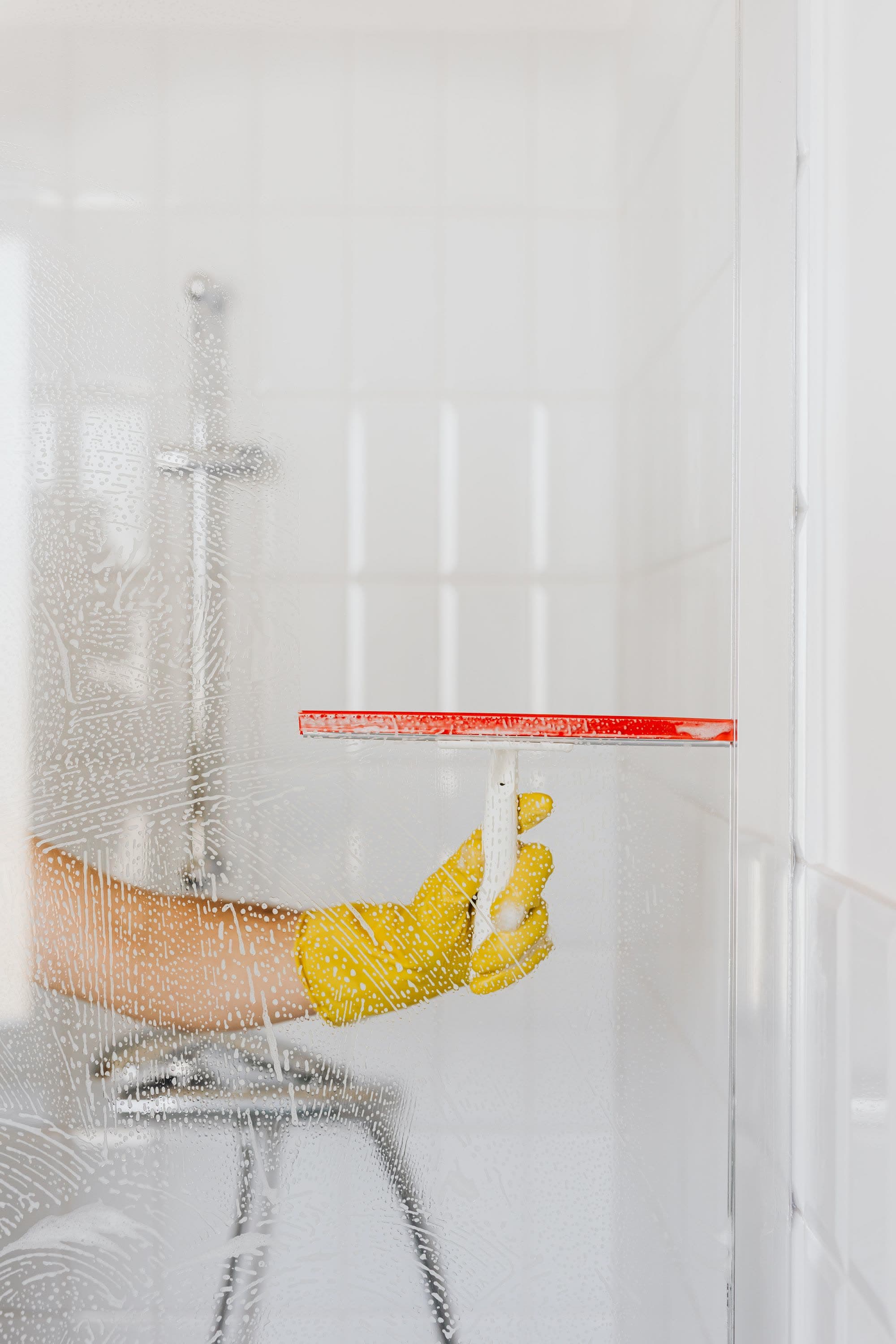 Hand i gul gummihandske drar en skrapa över en duschvägg i glas